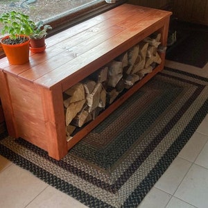 May include: A wooden firewood storage bench with a reddish-brown finish. The bench is rectangular with an open front, filled with stacked firewood. Two small potted plants sit on top. A braided rug is beneath the bench.