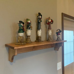 Puede incluir: Una estantería de madera con cinco figuras de muñeco de nieve. La estantería está teñida de un marrón oscuro y tiene un borde decorativo. Los muñecos de nieve son todos de diferentes colores y diseños, y algunos sostienen objetos como una linterna o una escoba.