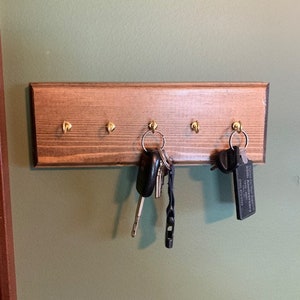 May include: A wooden key rack with five hooks, painted in a dark brown stain.  The rack has a set of keys hanging from one of the hooks.  The keys are silver and black with a black key fob that says "South Pointe Pharmacy".