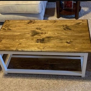 Puede incluir: Una mesa de centro de madera blanca y marrón con un estante inferior. La superficie de la mesa está hecha de madera teñida y la base está pintada de blanco.