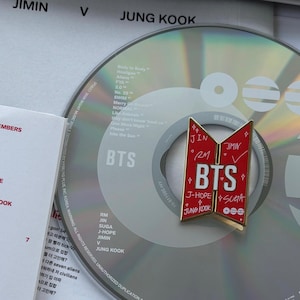 Op de afbeelding: Een BTS-album met een rode en witte pin met het BTS-logo en namen van leden. De cd is zilverkleurig met songtitels. Een wit papier vermeldt de leden: RM, Jin, Suga, J-Hope, Jimin, V en Jung Kook.