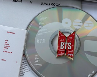 May include: A BTS album with a red and white pin featuring the BTS logo and member names. The CD is silver with song titles. A white paper lists members: RM, Jin, Suga, J-Hope, Jimin, V, and Jung Kook.