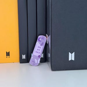 May include: A light purple bookmark with the text "bts are back" and a smiley face, moon, and butterfly design. The bookmark is leaning against a stack of black books and a yellow book with the word "Cream" on the spine.