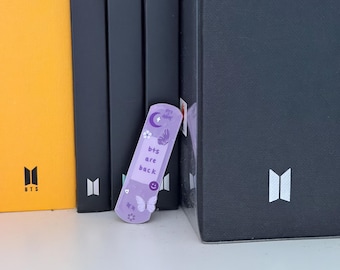 May include: A light purple bookmark with the text "bts are back" and a smiley face, moon, and butterfly design. The bookmark is leaning against a stack of black books and a yellow book with the word "Cream" on the spine.