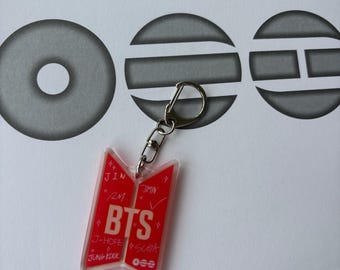 May include: A red acrylic keychain shaped like the BTS logo, featuring white text that reads BTS, Jin, RM, J-Hope, Jung Kook, Jimin, and Suga. The keychain includes a silver-colored metal lobster clasp and small star accents printed on the surface.