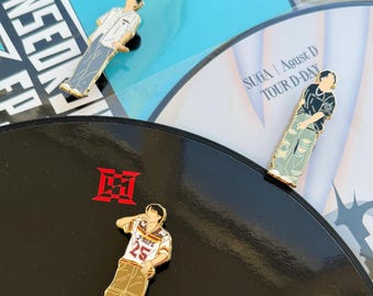 May include: Enamel pins featuring stylized figures in various outfits. One pin depicts a figure in a striped shirt and gray pants. Another shows a figure in a black shirt and ripped jeans. A third pin shows a figure in a jersey with the number 25.