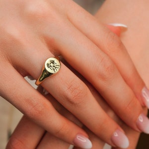 May include: A gold signet ring with a detailed design on the top. The ring is worn on the index finger of a hand.