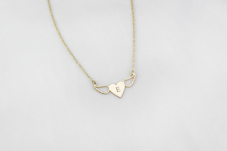 Op de afbeelding: Een gouden ketting met een hartvormige hanger met een enkele letter 'E' en vleugels.