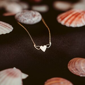 Op de afbeelding: Een gouden ketting met een hartvormige hanger en vleugels. De ketting ligt op een zwarte achtergrond met schelpen.