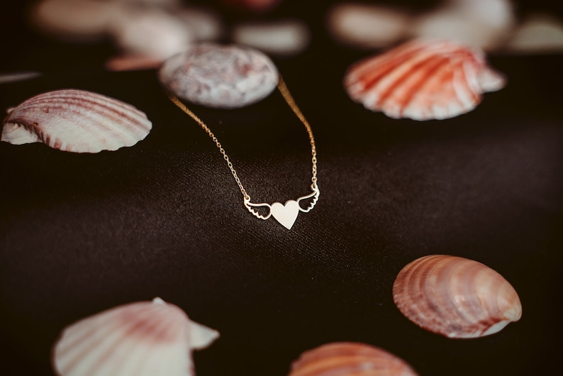 Op de afbeelding: Een gouden ketting met een hartvormige hanger en vleugels. De ketting ligt op een zwarte achtergrond met schelpen.