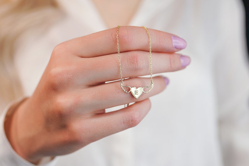 Op de afbeelding: Een gouden ketting met een hartvormige hanger met een enkele initiaal "E" en kleine vleugels aan beide kanten.