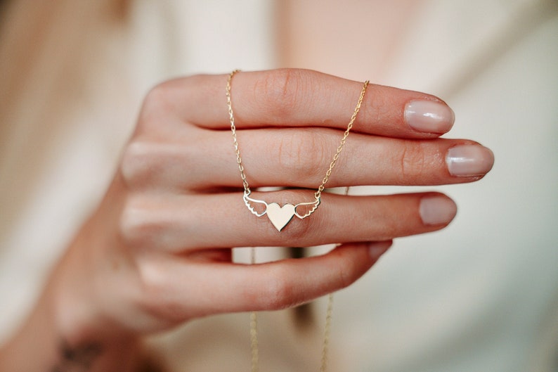 Op de afbeelding: Een gouden ketting met een hartvormige hanger met delicate vleugels. Het hart is een eenvoudig, minimalistisch ontwerp.