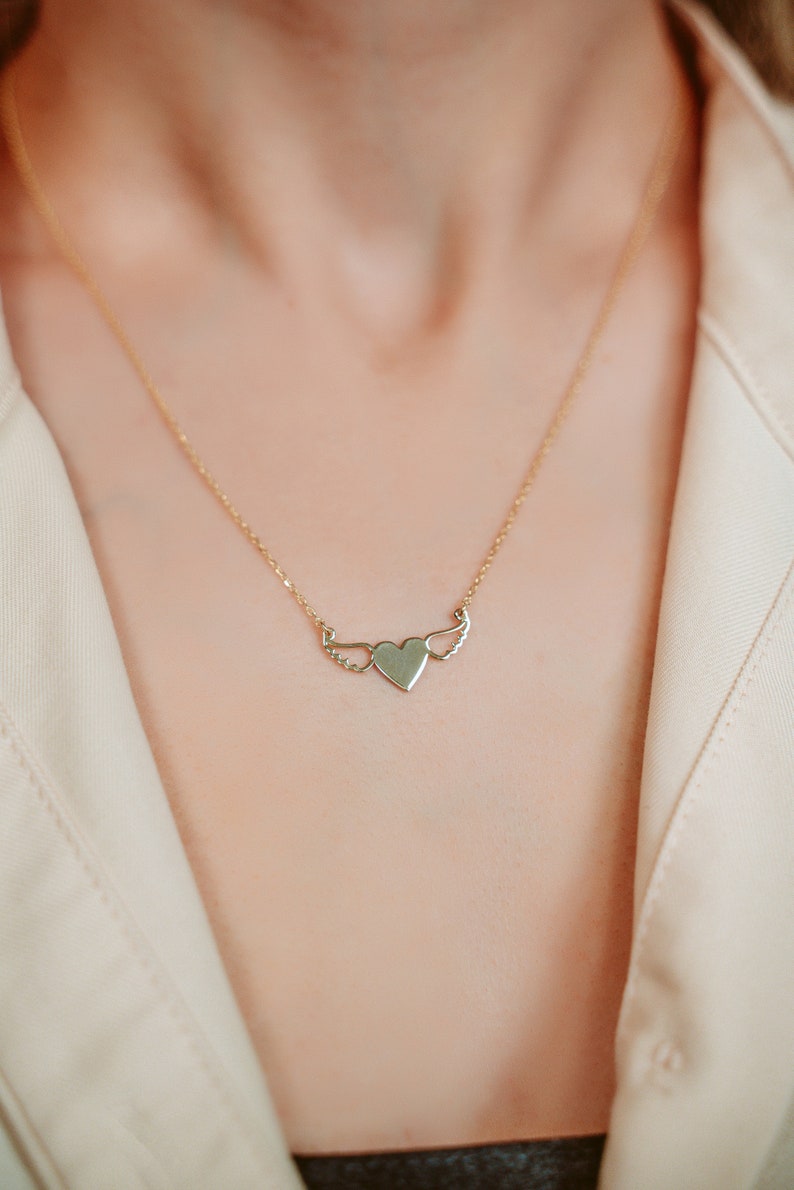 Op de afbeelding: Een gouden ketting met een hartvormige hanger met delicate vleugels. Het hart is glad en de vleugels zijn gedetailleerd.
