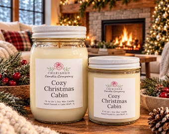 May include: Two glass jars of soy wax candles with white lids and labels reading Cherished Candle Company Cozy Christmas Cabin. The larger jar is 16 oz and the smaller is 8 oz. Both feature a pink rose logo and text detailing the contents.
