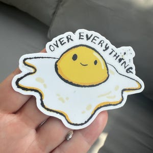 May include: A white sticker with a cartoon fried egg design. The egg has a yellow yolk with a smiling face and the words "OVER EVERYTHING" in a circular pattern around the egg.