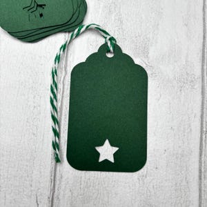 May include: Dark green gift tags with a white star cutout and scalloped top edge. Each tag is attached to a green and white twine string. A stack of similar tags is visible in the background.
