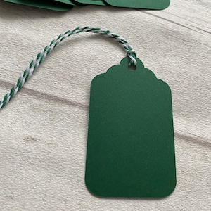 May include: Dark green gift tags with a scalloped top edge and a small hole for stringing. Each tag is attached to a white and green twisted twine. The tags are arranged on a light-coloured wooden surface.