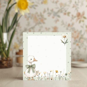 Könnte beinhalten: Quadratischer Notizblock mit einer Aquarellillustration einer weißen Ente mit grüner Schleife. Der Notizblock hat einen hellgrünen Rand mit kleinen Sternen und einer Gänseblümchen. Der untere Teil zeigt ein Feld mit Wildblumen.