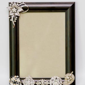 May include: A black picture frame with a beige mat and a decorative border of rhinestones. The frame is decorated with five brooches, including a floral brooch in the top corner.