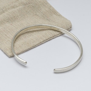 Può includere: Un bracciale a polsino in argento dal design minimalista, appoggiato su un sacchetto di lino beige. Il bracciale ha una superficie liscia e lucida e un design semplice e aperto. Il sacchetto offre uno sfondo neutro.