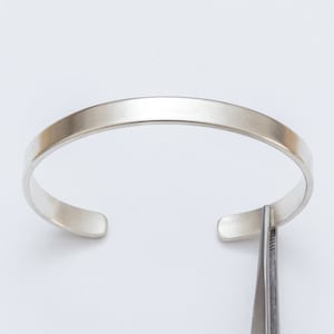 Może przedstawiać: Srebrna bransoletka typu bangle o gładkim, polerowanym wykończeniu. Bransoletka jest otwarta i ma lekko zakrzywiony kształt.