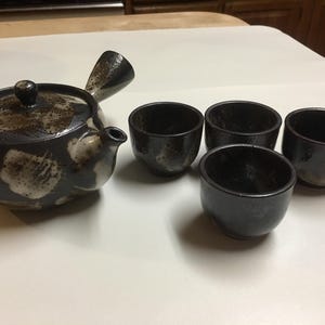 Op de afbeelding: Een keramische theeset met een theepot met een zijhandvat en vier bijpassende theekopjes. De set is donkerbruin met witte bloemaccenten. De theekopjes en de theepot zijn op een wit oppervlak geplaatst.