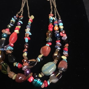 May include: Multi-strand necklace featuring a variety of colorful beads and stones. The necklace includes red, blue, green, and brown stones, with a gold-colored chain.