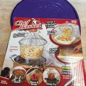 Könnte beinhalten: Eine rot-weiße Schachtel für den Chef Basket Deluxe, ein 12-in-1-Küchenwerkzeug. Die Schachtel zeigt den Korb beim Dämpfen, Spülen, Abseihen und Frittieren. Der Korb ist aus massivem Stahl und spülmaschinenfest.