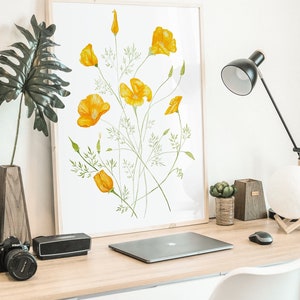 May include: A framed print of a watercolour painting of yellow California poppies on a white background. The print is hanging on a white wall above a desk with a laptop, a camera, and a lamp.