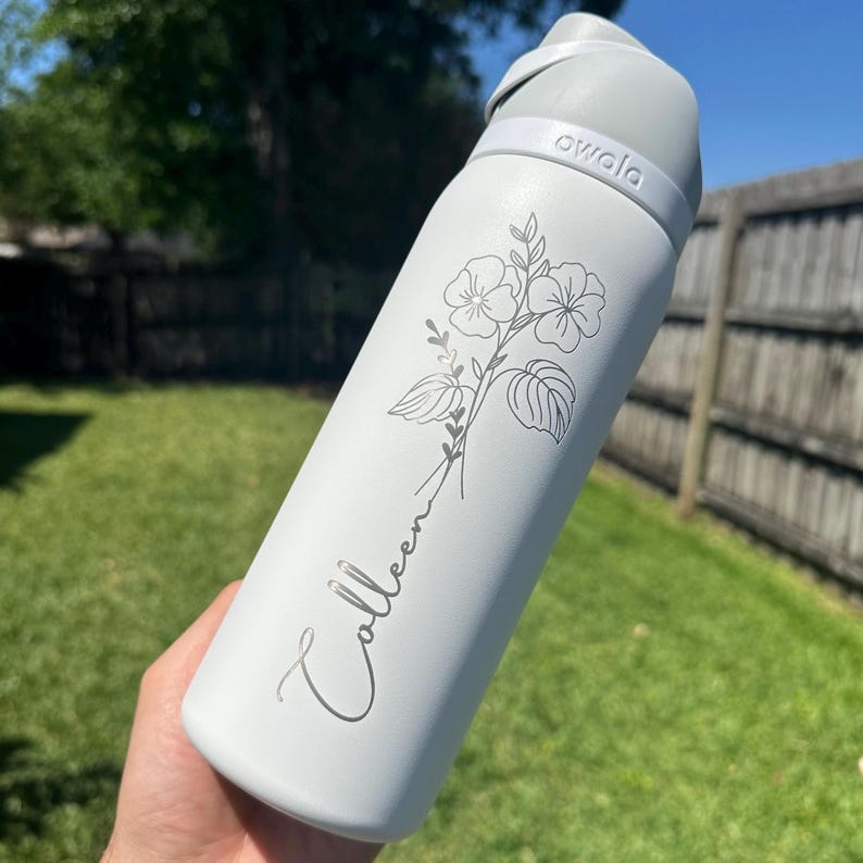 Puede incluir: Una botella de agua Owala blanca con tapa y asa grises. La botella est&aacute; grabada con un dise&ntilde;o floral y el nombre "Colleen" en cursiva. La botella se sostiene al aire libre frente a un c&eacute;sped verde y una valla de madera.