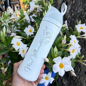 Puede incluir: Una botella de agua blanca con tapa y asa grises. La botella tiene un dise&ntilde;o floral y el nombre "Mrs. Williams" en cursiva. La marca "owala" est&aacute; impresa en la tapa.