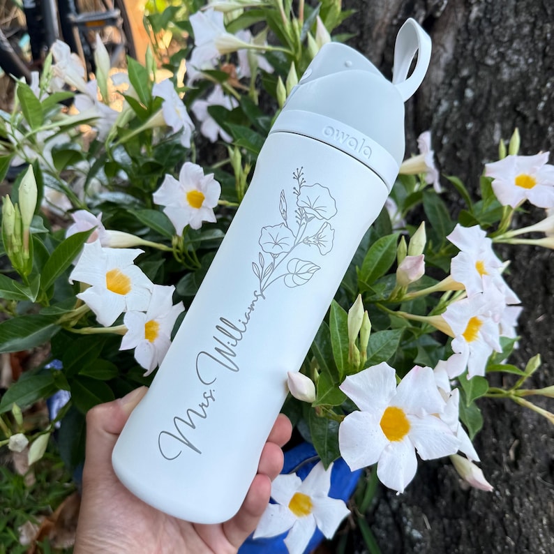 Puede incluir: Una botella de agua blanca con tapa y asa grises. La botella tiene un dise&ntilde;o floral y el nombre "Mrs. Williams" en cursiva. La marca "owala" est&aacute; impresa en la tapa.
