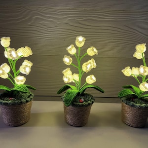 May include: Three decorative, illuminated lily-of-the-valley plants in woven pots. Each plant features glowing white flowers, green leaves, and a green stem. The pots are filled with green moss and have a small ladybug.