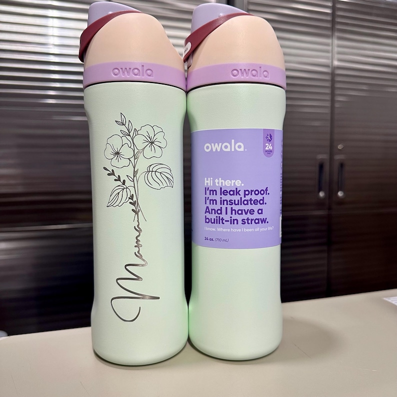 Puede incluir: Dos botellas de agua Owala. Una es verde claro con un dise&ntilde;o floral y la palabra "Mama" en cursiva. La otra es verde claro con una etiqueta morada que dice "Hi there. I'm leak proof. I'm insulated. And I have a built-in straw."