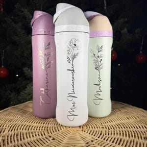 Puede incluir: Tres botellas de agua aisladas en tonos lavanda, blanco y verde menta. Cada botella presenta un dise&ntilde;o floral y texto personalizado. Las botellas tienen una tapa abatible y se exhiben sobre una superficie tejida.