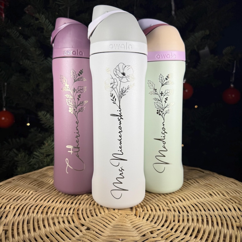 Puede incluir: Tres botellas de agua aisladas en tonos lavanda, blanco y verde menta. Cada botella presenta un dise&ntilde;o floral y texto personalizado. Las botellas tienen una tapa abatible y se exhiben sobre una superficie tejida.