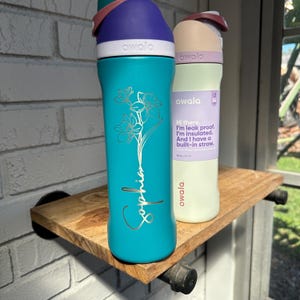 Puede incluir: Dos botellas de agua aisladas con tapas abatibles. Una es de color turquesa con un dise&ntilde;o floral y el nombre "Sophia". La otra es verde p&aacute;lido con una etiqueta morada. Ambas botellas tienen la marca "owala".