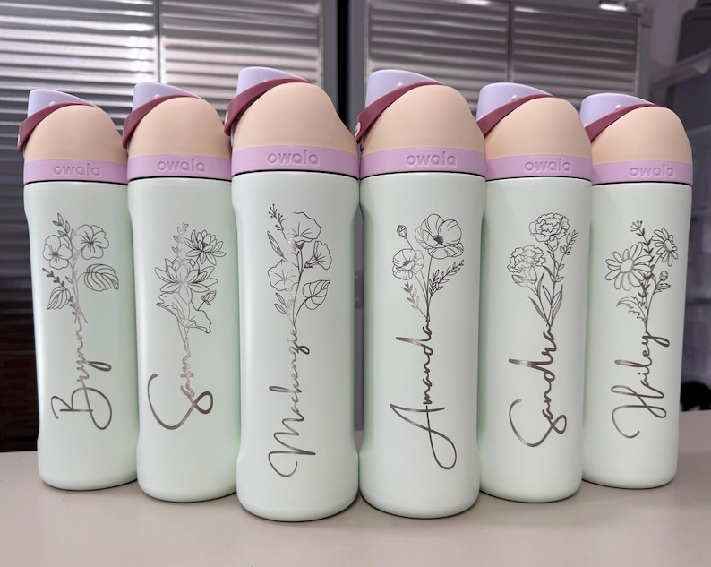 Puede incluir: Seis botellas de agua Owala en fila, cada una con un nombre diferente en escritura cursiva y un dise&ntilde;o floral. Las botellas son de color verde claro con una tapa rosa y beige. Los nombres son Brynne, Sophie, Mackenzie, Amanda, Sandra y Hailey.
