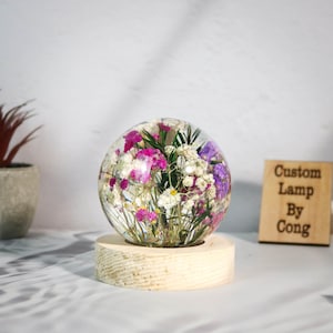 May include: A clear glass sphere encasing colorful dried flowers, including pink, white, and purple blooms. The sphere sits atop a light-colored wooden base. A wooden sign with the text "Custom Lamp By Cong" is visible.