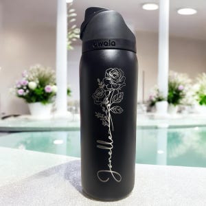 Puede incluir: Botella de agua aislada negra con un dise&ntilde;o floral de rosas y el nombre "Janelle" en cursiva. La botella tiene una tapa abatible negra y el nombre de la marca "owala" est&aacute; impreso en la tapa.