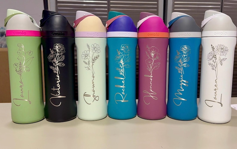 Puede incluir: Una colecci&oacute;n de botellas de agua aisladas en varios colores, incluyendo verde, negro, menta, turquesa, magenta, azul y blanco. Cada botella presenta un dise&ntilde;o floral y un nombre en fuente cursiva. Las botellas tienen una tapa abatible.