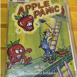 Puede incluir: Un cartucho de juego Atari vintage para "Apple Panic". La portada presenta una manzana de dibujos animados con boca, un granjero y el título del juego en letras rojas. El fondo es verde con una escalera y otros elementos del juego.
