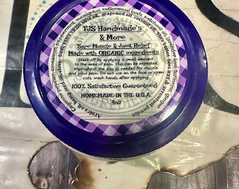 以下が含まれることがあります： 白いラベルに「TJS Handmade's & More Sore Muscle & Joint Relief Made with ORGANIC ingredients 100% Satisfaction Guaranteed HOMEMADE IN THE U.S.A. 4oz」と書かれた紫色の瓶。瓶は木製の表面の上に置かれています。
