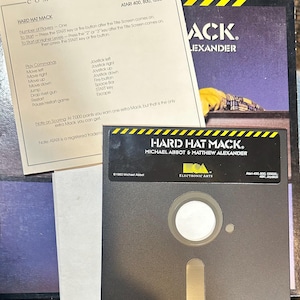 May include: A black floppy disk with a white label and a circular hole in the center. The label reads "Hard Hat Mack. Michael Abbot & Matthew Alexander. Electronic Arts. Atari 400, 800, 1200. 4SK Joystick."