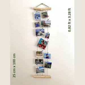 Può includere: Un espositore fotografico in legno con più foto attaccate a delle corde. Il display misura 25 cm x 100 cm e 0,82 ft x 3,28 ft. Le foto sono di varie dimensioni e raffigurano persone e paesaggi. Il display è appeso a una parete bianca.