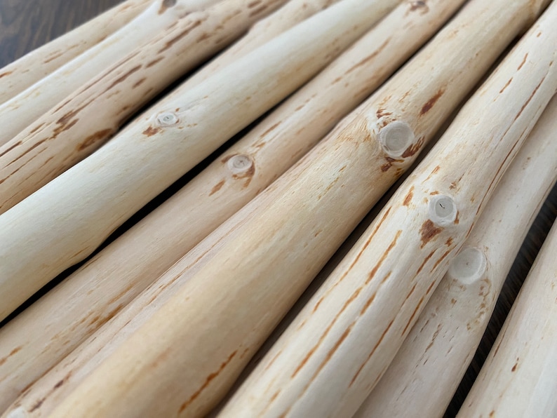 Pu&ograve; includere: Primo piano di diversi pali di legno color chiaro, possibilmente rami, disposti diagonalmente. I pali hanno un aspetto naturale, non rifinito, con nodi visibili e sottili venature. Il legno ha una tonalit&agrave; chiara e neutra, con striature marrone scuro.