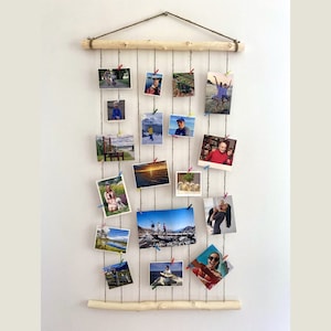 Op de afbeelding: Een rustieke fotodisplay gemaakt van natuurlijk hout en touw, hangend aan een witte muur. De display bevat meerdere foto's die met kleurrijke wasknijpers zijn vastgezet. De foto's tonen verschillende scènes en mensen, waardoor een gepersonaliseerde wanddecoratie ontstaat.