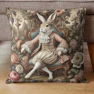 May include: Decorative throw pillow featuring a regal rabbit in antique attire seated on a blue armchair. The rabbit is surrounded by a garden of roses and other flowers, with a vintage aesthetic.