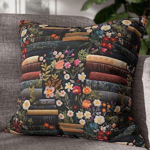 May include: Square throw pillow with a floral and book design. The pillow features a dark background with stacks of books and colorful flowers in shades of pink, red, white, and yellow. The pillow is on a gray sofa.