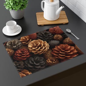 May include: A rectangular placemat featuring a detailed pattern of various-sized pine cones in shades of brown, tan, and black. The placemat is on a dark surface, with a white teacup and saucer, a small potted plant, and a teapot in the background.